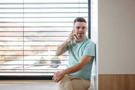 Téléchargez les photos : Portrait de jeune homme trisomique assis par la fenêtre, faisant téléphone avec smartphone. - en image libre de droit