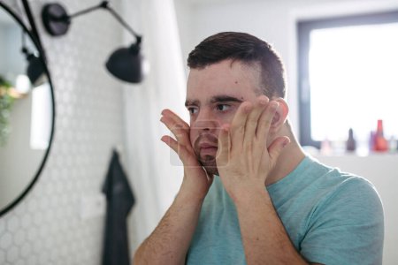 Téléchargez les photos : Vue arrière du jeune homme avec le syndrome du duvet dans la salle de bain, tenant ses joues, va se raser la barbe. - en image libre de droit