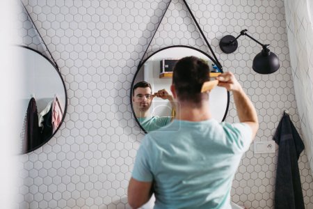Téléchargez les photos : Vue arrière du jeune homme avec le syndrome du duvet dans la salle de bain, peigner ses cheveux. - en image libre de droit