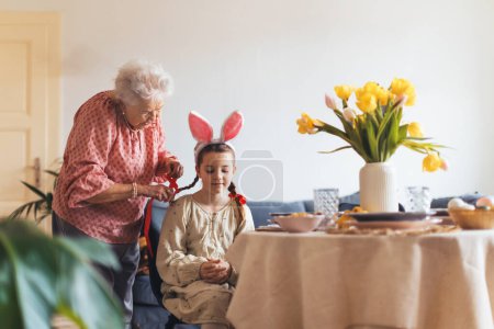 Grandmother braiding hair of granddaughter, putting ribbon ob braid. Prepairing for easter lunch.. Concept of family easter holidays.