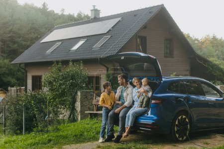 Téléchargez les photos : Famille avec voiture électrique debout devant leur maison avec des panneaux solaires sur le toit. Énergie solaire et mode de vie durable de la jeune famille. Concept d'énergie verte et d'avenir durable pour la prochaine - en image libre de droit