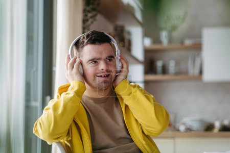 Photo for Young man with down syndrome listening his music via headphones. Sitting in the kitchen, wireless headphones on head. - Royalty Free Image