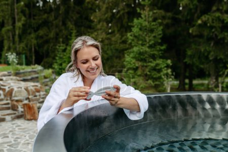 Téléchargez les photos : Young woman in bathrobe, checking temperature with thermometer, ready for home spa procedure in hot tub outdoors. Wellness, body care, hygiene concept. - en image libre de droit