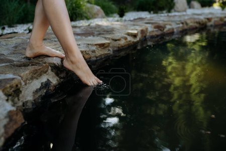Photo for An unrecognizable young woman is dipping her foot in cool water of pond, refreshing and hardening concept. - Royalty Free Image