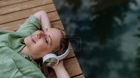 Foto de Vista superior de la mujer relajada con auriculares escuchando música sentada en un muelle junto a un lago natural en verano - Imagen libre de derechos