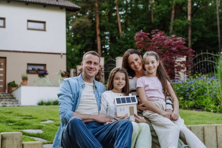 Happy family holding model of house with solar panels on roof and wind turbine model. Alternative, renewable, green energy and sustainable lifestyle concept.