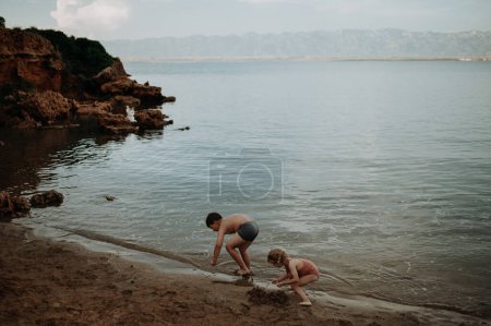 Foto de Hermano ayudando a hermana seraching para conchas en arena mojada. Niña en traje de baño jugando en la playa, agachada en el agua. - Imagen libre de derechos