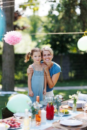 Family portrait at outdoor summer garden party. Aunt with niece standing by table with food and posing for photo.