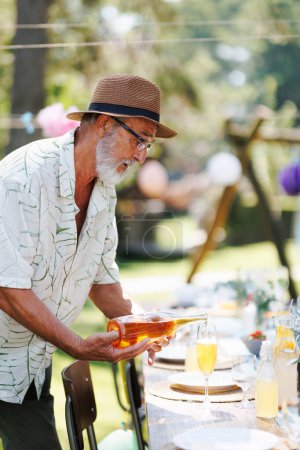 Foto de Anciano guapo hombre preparando refrescos para una fiesta de jardín de verano, vertiendo vino en un vaso. - Imagen libre de derechos