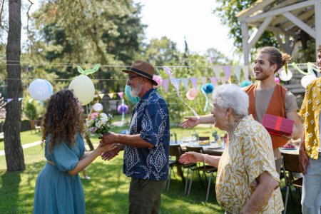 Grandfather gifts a wrapped present to granddaughter as surprise at birthday party. Friends and family reunite at garden party.