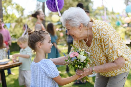 Photo for Garden birthday party for senior lady. Beautiful senior birthday woman receiving gift from granddaughter, hugging. - Royalty Free Image