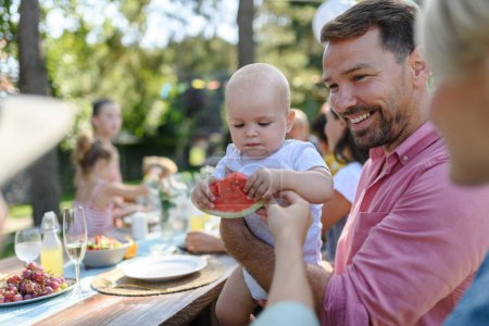Photo for Cute baby eating watermelon at a summer garden party. Handsome father holding the baby boy in his arms and smiling. - Royalty Free Image
