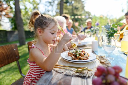 Téléchargez les photos : Portrait d'une belle petite fille assise à une table mangeant des aliments grillés à l'extérieur. Fille à la fête de jardin familiale. - en image libre de droit