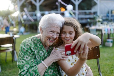 Photo for Young girl taking selfie photo with elderly grandmother at a garden party. Love and closeness between grandparent and grandchild. - Royalty Free Image