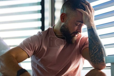 Photo for Anxious, depressed man in pink t-shirt with tattoo on arms, closed eyes, holding head in hand. - Royalty Free Image