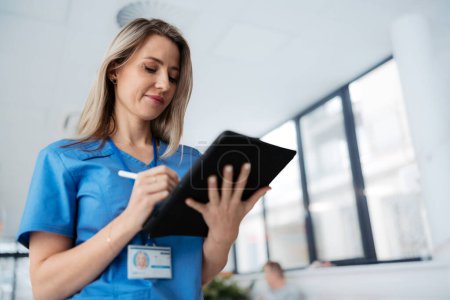 Photo for Portrait of confident female doctor standing in Hospital corridor. Beautiful nurse wearing blue scrubs, holding clipboard standing in modern private clinic, low angle photography. - Royalty Free Image