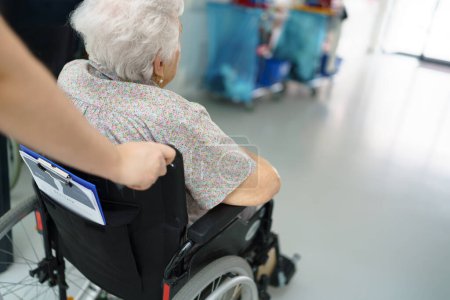 Close up of nurse pushing senior patient in a wheelchair across hospital corridor, hall. Emotional support for elderly woman.