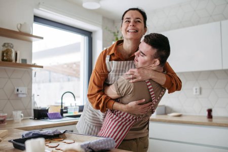Young man with Down syndrome baking cookies, sweets with his mother at home, hugging her. Daily routine for man with Down syndrome. Concept of mothers day.
