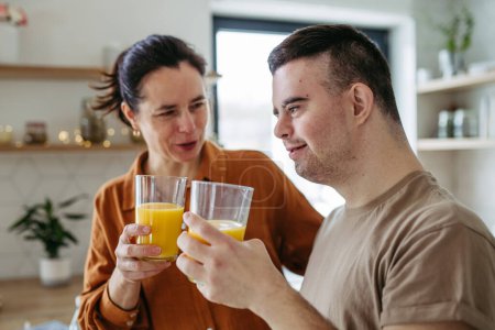 Photo for Portrait of young man with Down syndrome with his mother at home, toasting with juice. Morning routine for man with Down syndrome genetic disorder. - Royalty Free Image