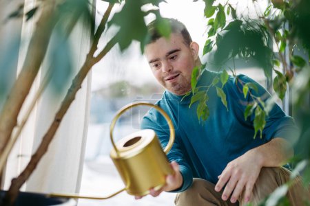 Young man with Down syndrome taking care of indoor plants, watering them, looking at the camera through handle of a metal watering can.