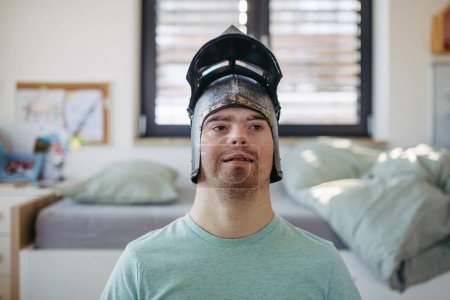 Portrait of happy smiling man with down syndrome at home, with knights helmet on head.