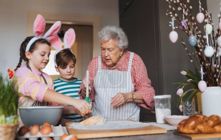 Téléchargez les photos : Grand-mère avec ses petits-enfants préparant des repas traditionnels de Pâques, pétrissant la pâte pour les petits pains croisés de Pâques. Transmission de recettes familiales, de coutumes et d'histoires. Concept de vacances de Pâques en famille. - en image libre de droit