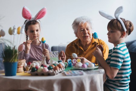 Foto de Abuela con niños pequeños decorando huevos de Pascua en casa. Tradición de pintar huevos con pincel y tinte de huevo de Pascua. Concepto de vacaciones familiares de Pascua. - Imagen libre de derechos