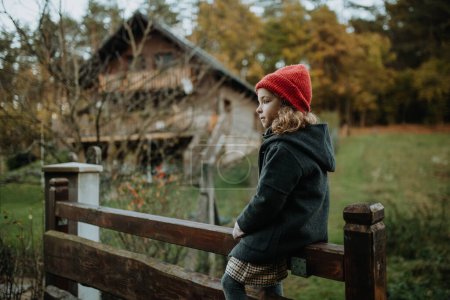Photo for Young girl in warm clothes sitting on aged wooden fence or wooden gate, looking at autumn nature. - Royalty Free Image