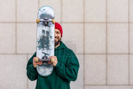 Foto de Retrato de skateboarder elegante con monopatín en frente de la cara, mirando a la cámara. Un joven patinando en la ciudad. Concepto de skate como deporte y estilo de vida. - Imagen libre de derechos