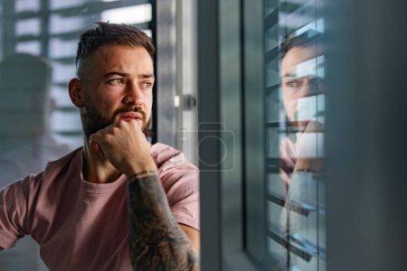 Photo for Handsome calm man in pink t-shirt with tattoo on arms looking through window. - Royalty Free Image