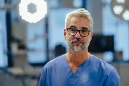 Téléchargez les photos : Portrait d'un médecin urgentiste confiant aux urgences de l'hôpital. Beau médecin en blouse bleue debout dans une clinique privée moderne, regardant la caméra. - en image libre de droit