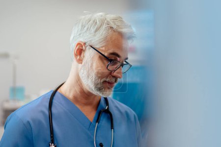 Portrait of confident ER doctor standing in hospital emergency room. Handsome doctor in blue scrubs with stethoscope, standing in modern private clinic.