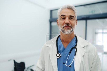 Portrait of confident mature doctor standing in Hospital corridor. Handsome doctor with gray hair wearing white coat, scrubs, stethoscope around neck standing in modern private clinic, looking at