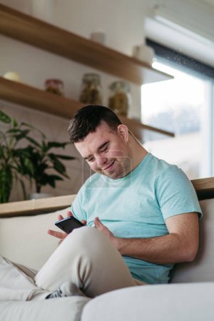 Téléchargez les photos : Portrait de jeune homme trisomique assis sur un canapé avec smartphone à la main et défilant. - en image libre de droit