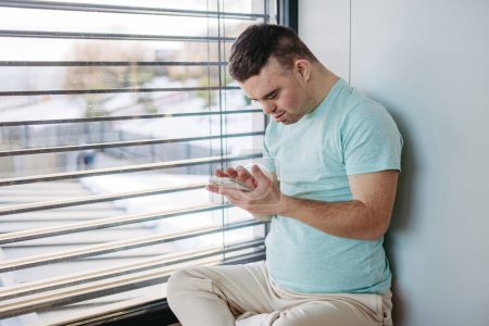 Téléchargez les photos : Portrait de jeune homme trisomique assis par la fenêtre avec smartphone à la main et défilement. - en image libre de droit