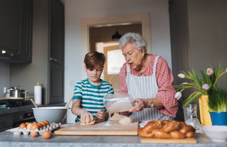 Téléchargez les photos : Grand-mère et petit-fils préparant les repas traditionnels de Pâques, pétrissant la pâte pour les petits pains croisés de Pâques. Transmission de recettes familiales, de coutumes et d'histoires. Concept de vacances de Pâques en famille. - en image libre de droit