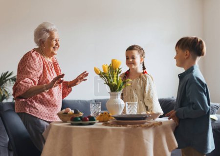 Photo for Grandmother with grandchildren setting table for traditional easter lunch. Recreating family traditions and customs. Concept of family easter holidays. Standing by table, praying. - Royalty Free Image