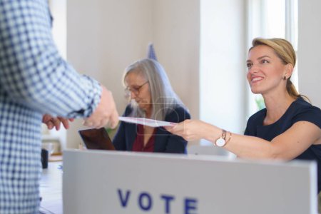 Foto de Mujer miembro de la comisión electoral en el lugar de votación el día de las elecciones, EE.UU. elecciones. Entrega de la boleta electoral al votante. - Imagen libre de derechos