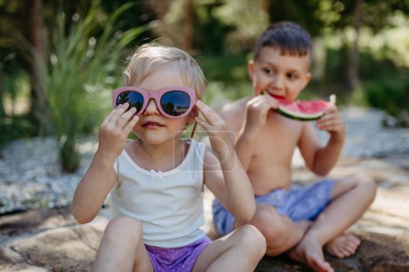 Téléchargez les photos : Little chidren sitting near a lake and eating watermelon on hot sunny day during summer vacation. - en image libre de droit