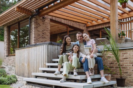 Téléchargez les photos : Happy family holding modèle de maison avec panneaux solaires sur le toit et le modèle d'éolienne. Concept d'énergies alternatives, renouvelables, vertes et de modes de vie durables. - en image libre de droit