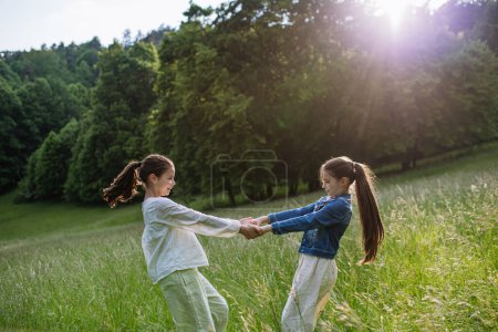 Two sisters playing at meadow in tall grass, running and dancing, having fun. Sisterly love and siblings relationship concept.