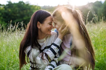 Photo for Beautiful mother with daughter, touching with foreheads and sitting in the grass at meadow. Concept of Mothers Day and maternal love. - Royalty Free Image