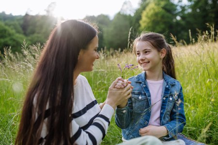 Photo for Beautiful mother with daughter, holding by hands and sitting in the grass at meadow. Concept of Mothers Day and maternal love. - Royalty Free Image