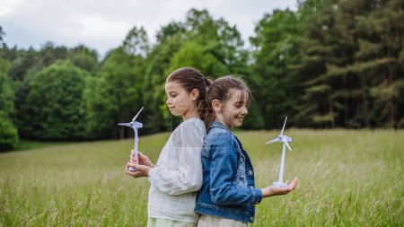 Photo for Two girls holding model wind turbine, standing on meadow, in nature. Alternative, renewable, green energy and sustainable lifestyle for future generations concept. - Royalty Free Image