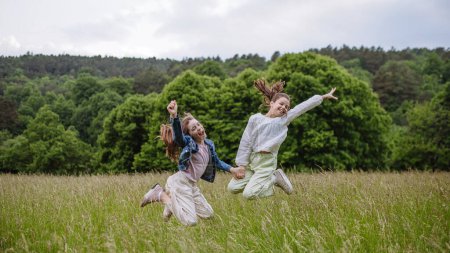 Two sisters playing at meadow in tall grass, running and jumping, having fun. Sisterly love and siblings relationship concept.