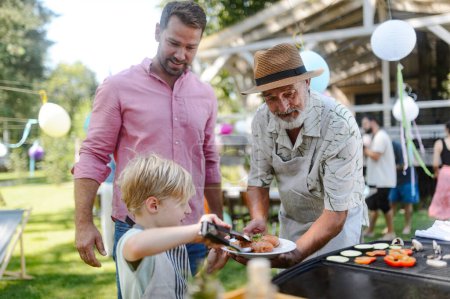 Téléchargez les photos : Père, grand-père et fils grillent ensemble lors d'une soirée barbecue dans le jardin. Trois générations d'hommes à la fête de jardin familial d'été. - en image libre de droit