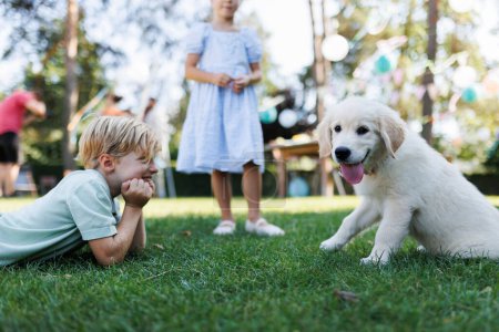 Foto de Niños jugando con un cachorro pequeño en una fiesta de jardín familiar. Retrato de niño tendido en la hierba mirando a Golden retriever cachorro. - Imagen libre de derechos