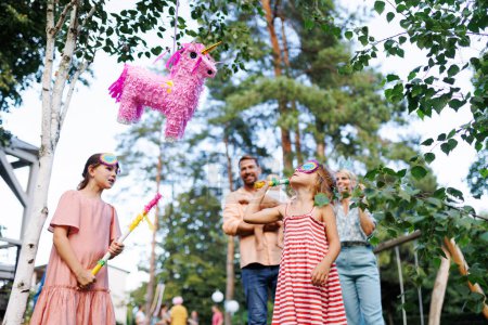 Foto de Dos chicas rompiendo, golpeando piñata rosa con un palo en la fiesta de cumpleaños. Los niños celebran el cumpleaños en la fiesta del jardín. Niños divirtiéndose y jugando. - Imagen libre de derechos