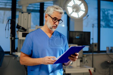 Portrait of confident ER doctor standing in hospital, emergency room. Handsome doctor in scrubs holding clipboard with test results, standing in modern private clinic, looking at camera.