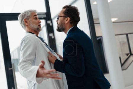 Photo for Violence against health workers. Patient is aggressive, have conflict with doctor in the hospital corridor. Visitor holding doctor by coat, screaming, threatening him. - Royalty Free Image